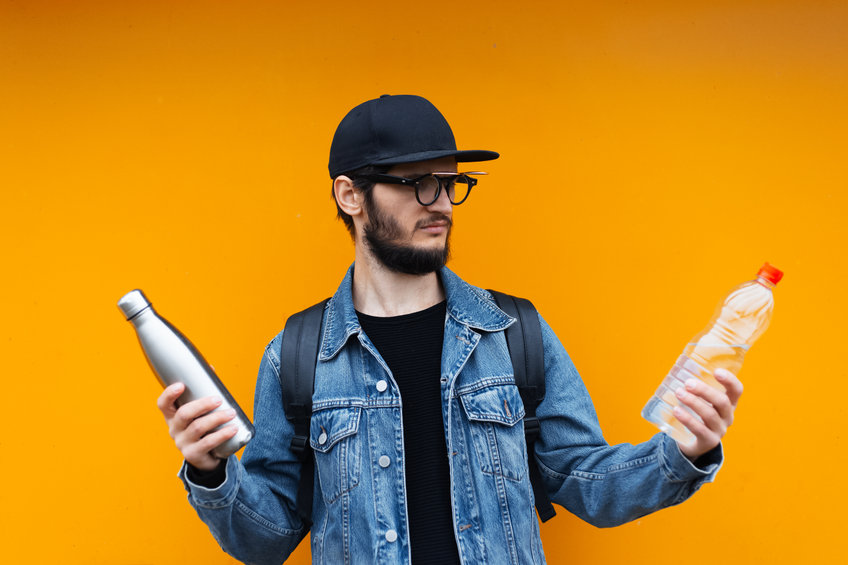Retrato de un joven hipster alegre, sosteniendo una botella termo ecológica de acero para agua y una botella de plástico, sobre fondo amarillo o naranja.  Di no al plástico.